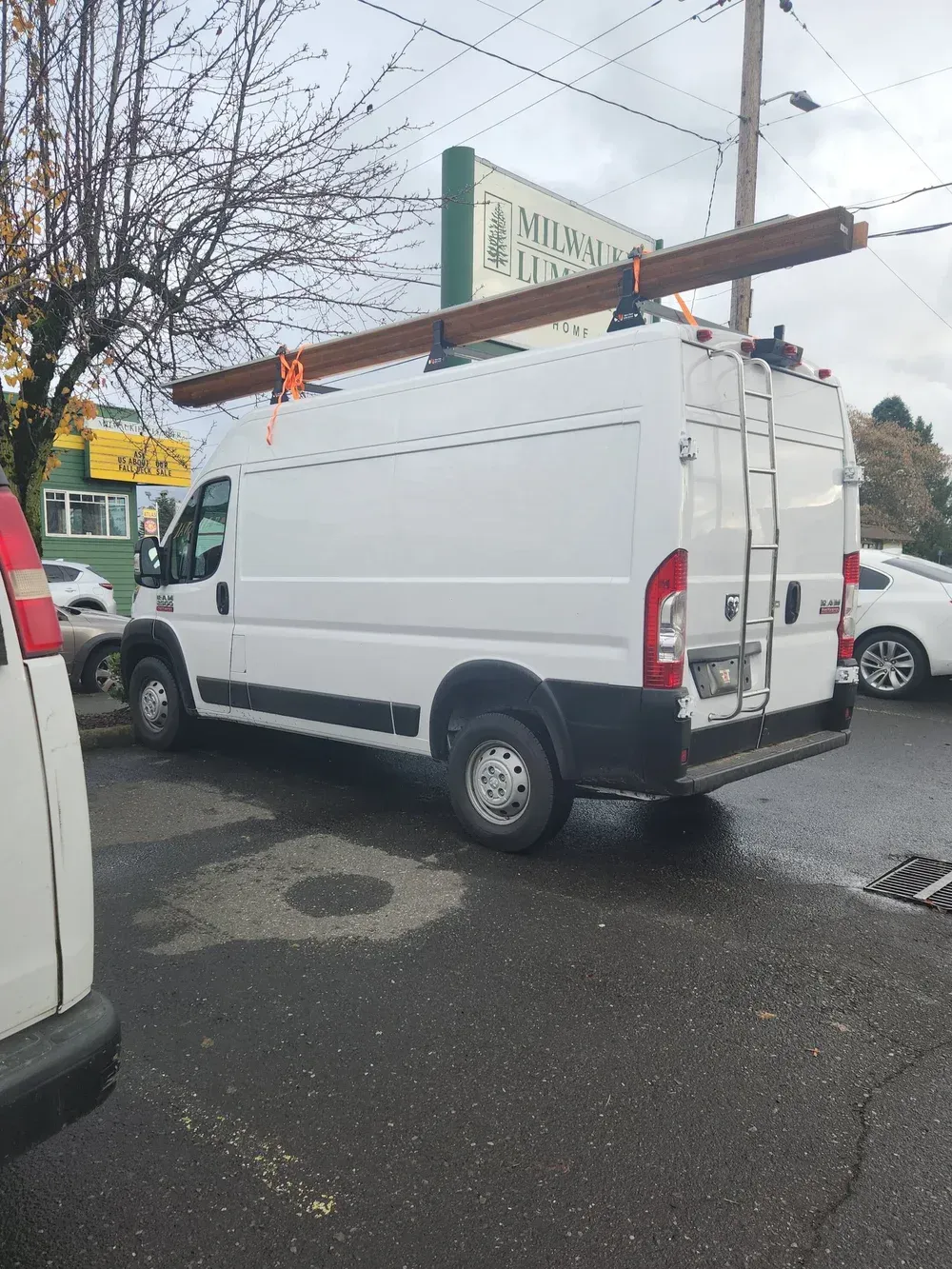 White work van with ladder and long beams secured on roof, parked on street.