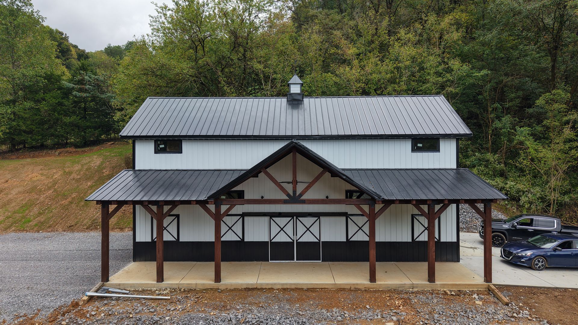 White barn with black roof and trim, wooden supports, and open doors, set against trees.