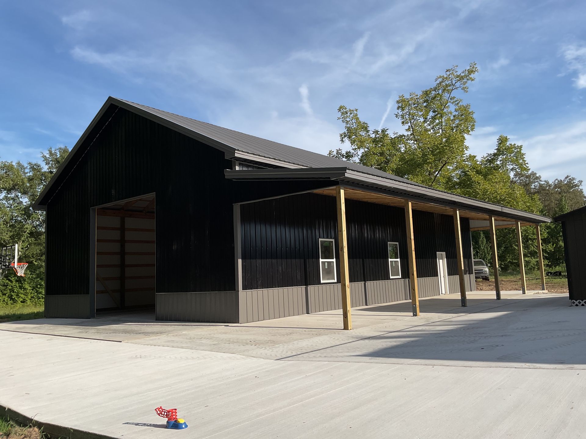 Black barn with a covered porch on a concrete pad under a blue sky, trees in the background.