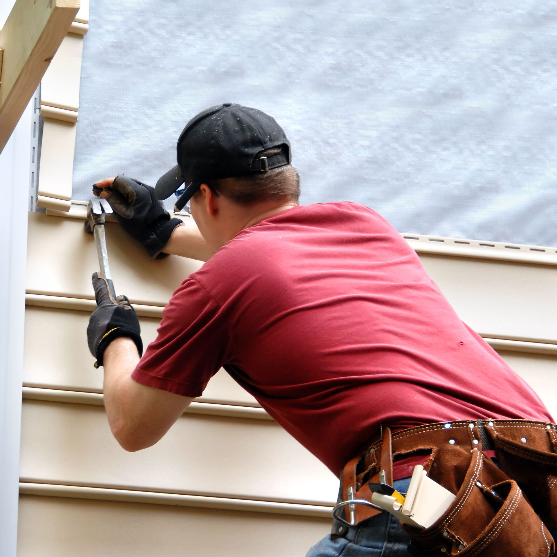 Worker using a hammer to install siding on a house exterior while wearing a tool belt and gloves