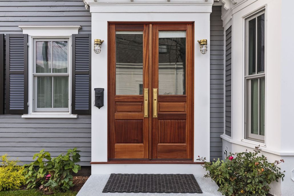 Wooden double front doors with white trim on a gray house, flanked by windows and small shrubs
