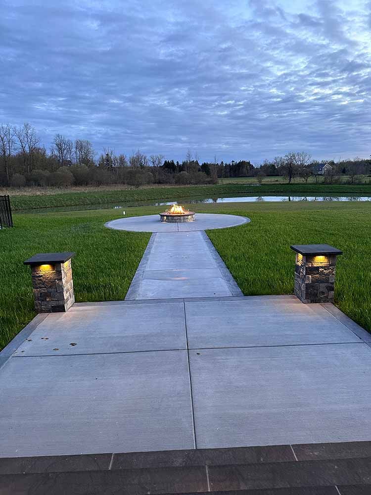 A concrete walkway leading to a fire pit in the middle of a grassy field.