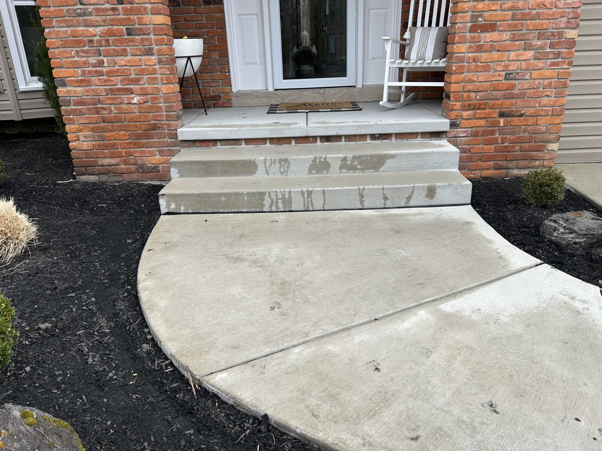 A brick house with a rocking chair in front of it and a concrete walkway leading to the front door.