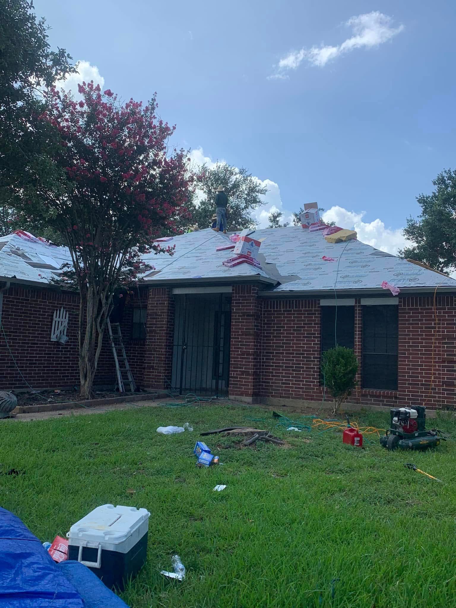 House with damaged roof, brick exterior, debris on lawn, ladder against tree, sunny day.