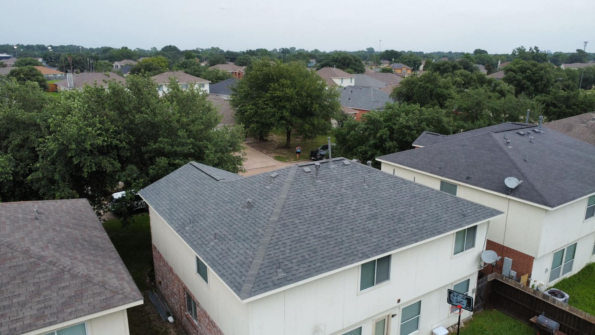 Aerial view of suburban houses with gray roofs and green trees under a cloudy sky.