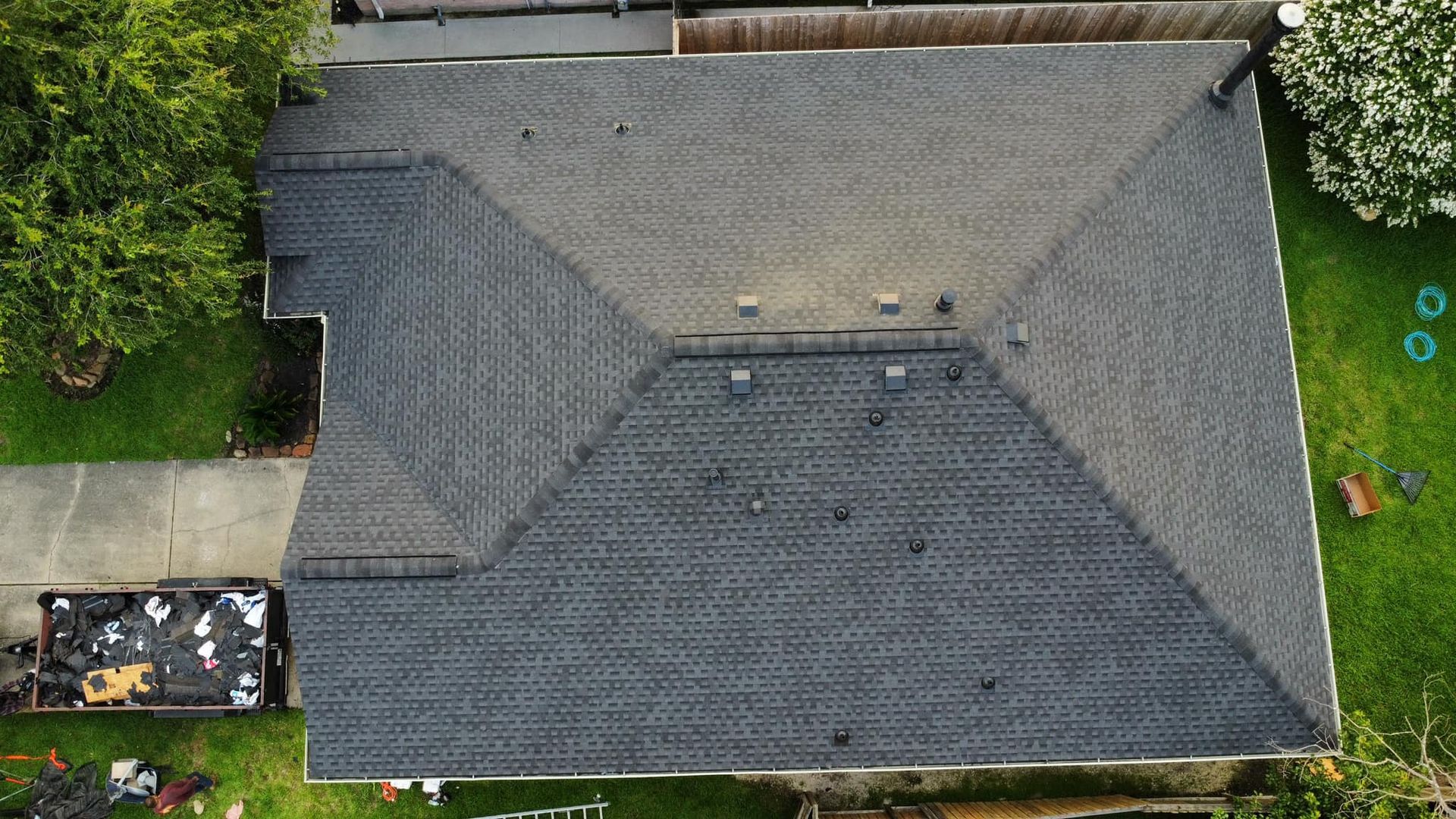 Overhead view of a dark gray asphalt shingle roof. A green lawn surrounds the house; a tree and driveway are visible.