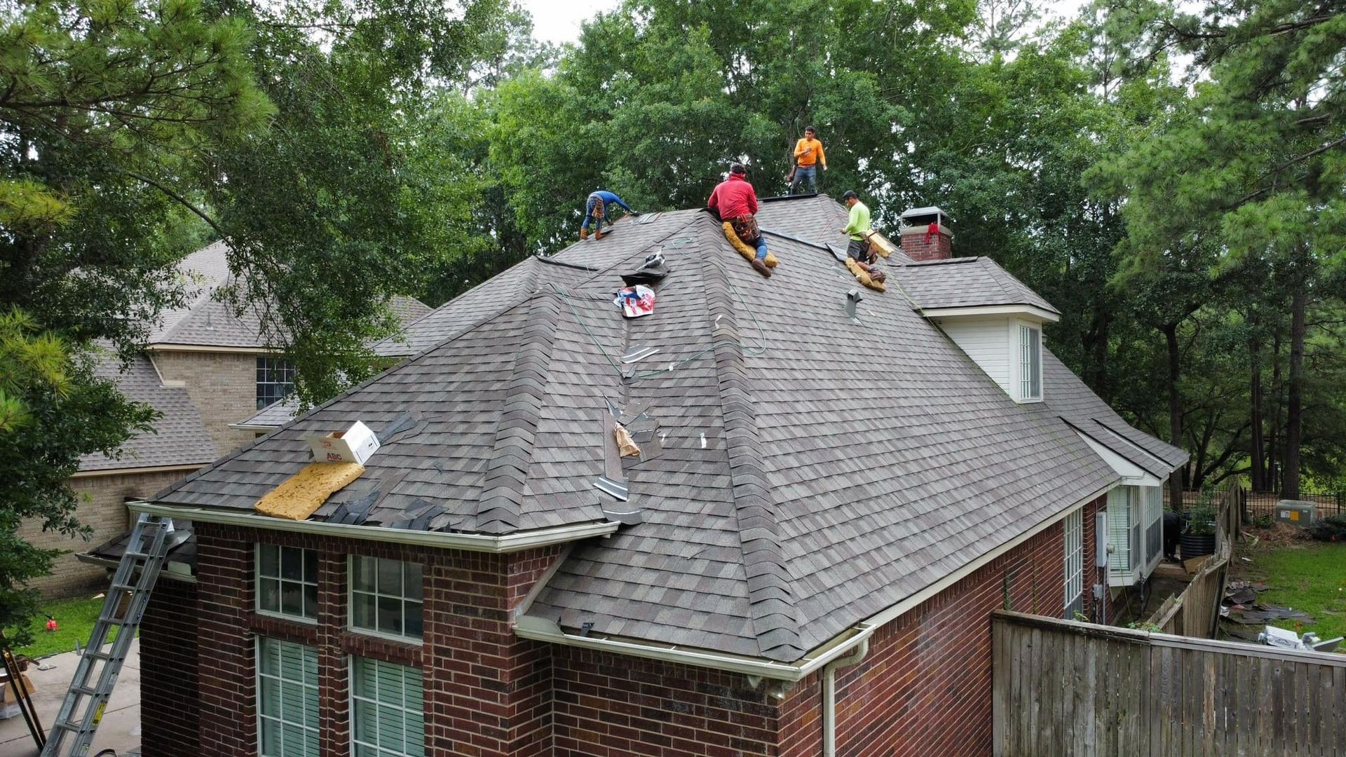 Roofers replacing shingles on a brick house roof; several workers on the roof, trees in the background.