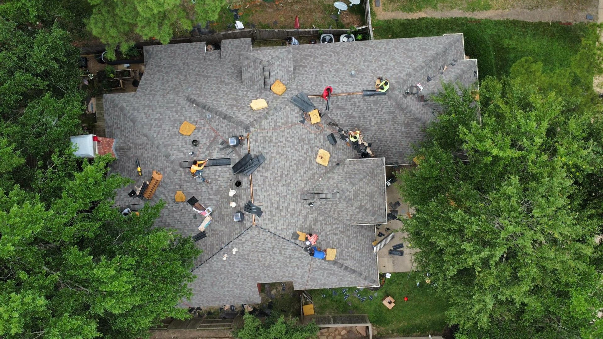 Overhead view of a house roof with workers and materials, likely undergoing repair or replacement. Trees surround the house.