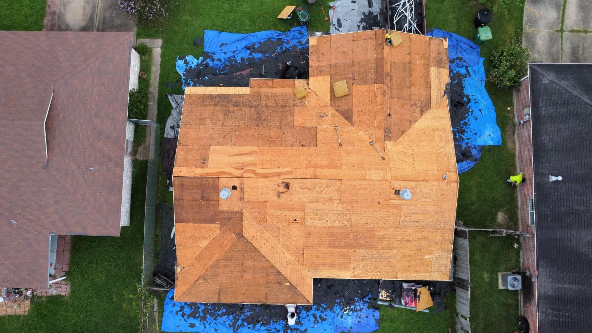 Overhead view of a house roof partially covered with new wood, with blue tarp and surrounding green lawn.
