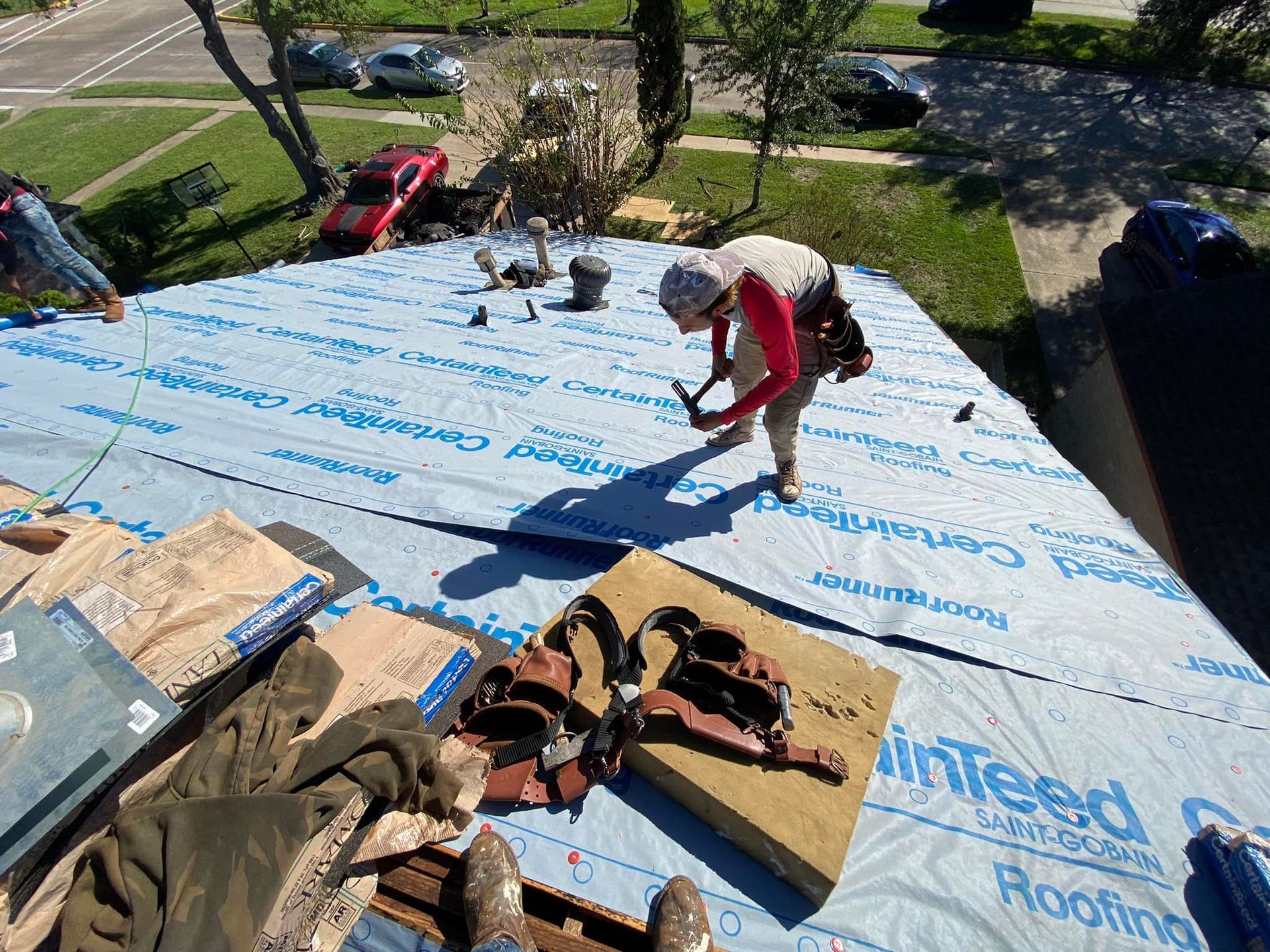 Roofer on a blue tarp-covered roof, hammering shingles in bright sunlight.