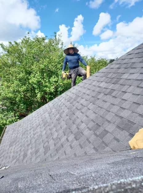 Roofer with straw hat on a shingled roof under a partly cloudy sky, holding materials.