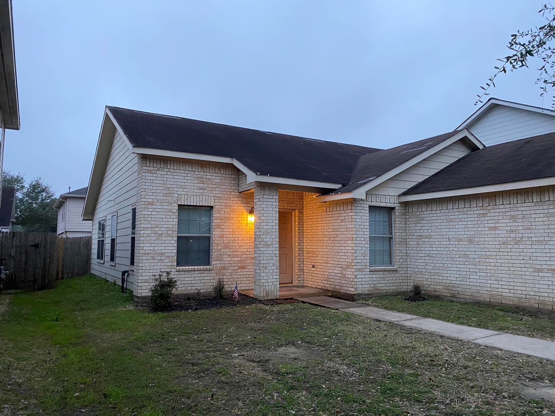 Brick house with a dark roof and a lit porch light on a cloudy day.