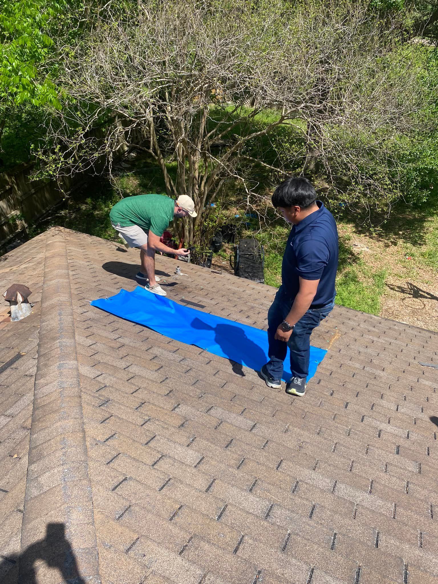 Two people on a rooftop with a blue mat. One bends over, the other stands. Sunlight. Trees in the background.