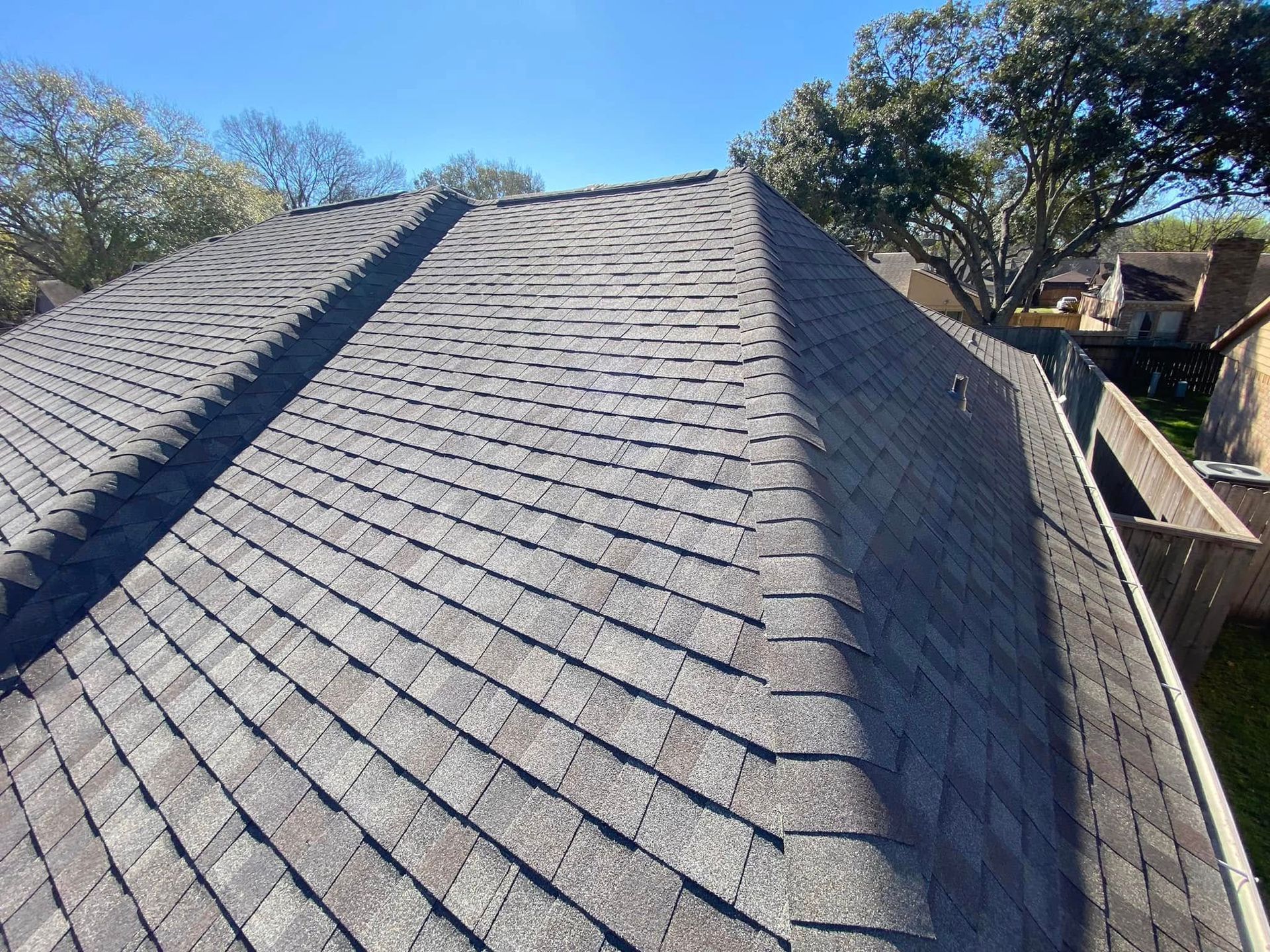Gray asphalt shingle roof on a house, angled view, sunny day. Trees in the background.