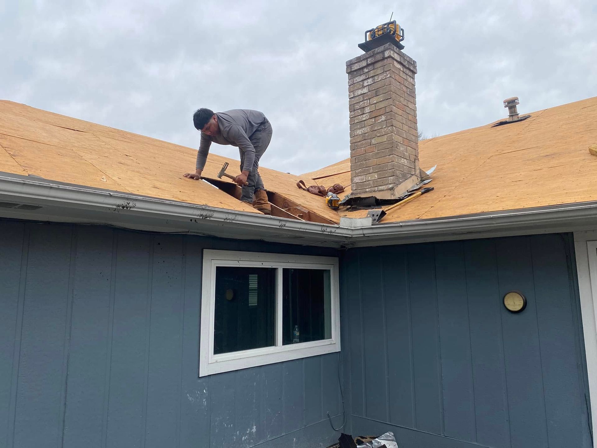 Person on a roof near a chimney, removing shingles. Blue house with a white window. Overcast sky.