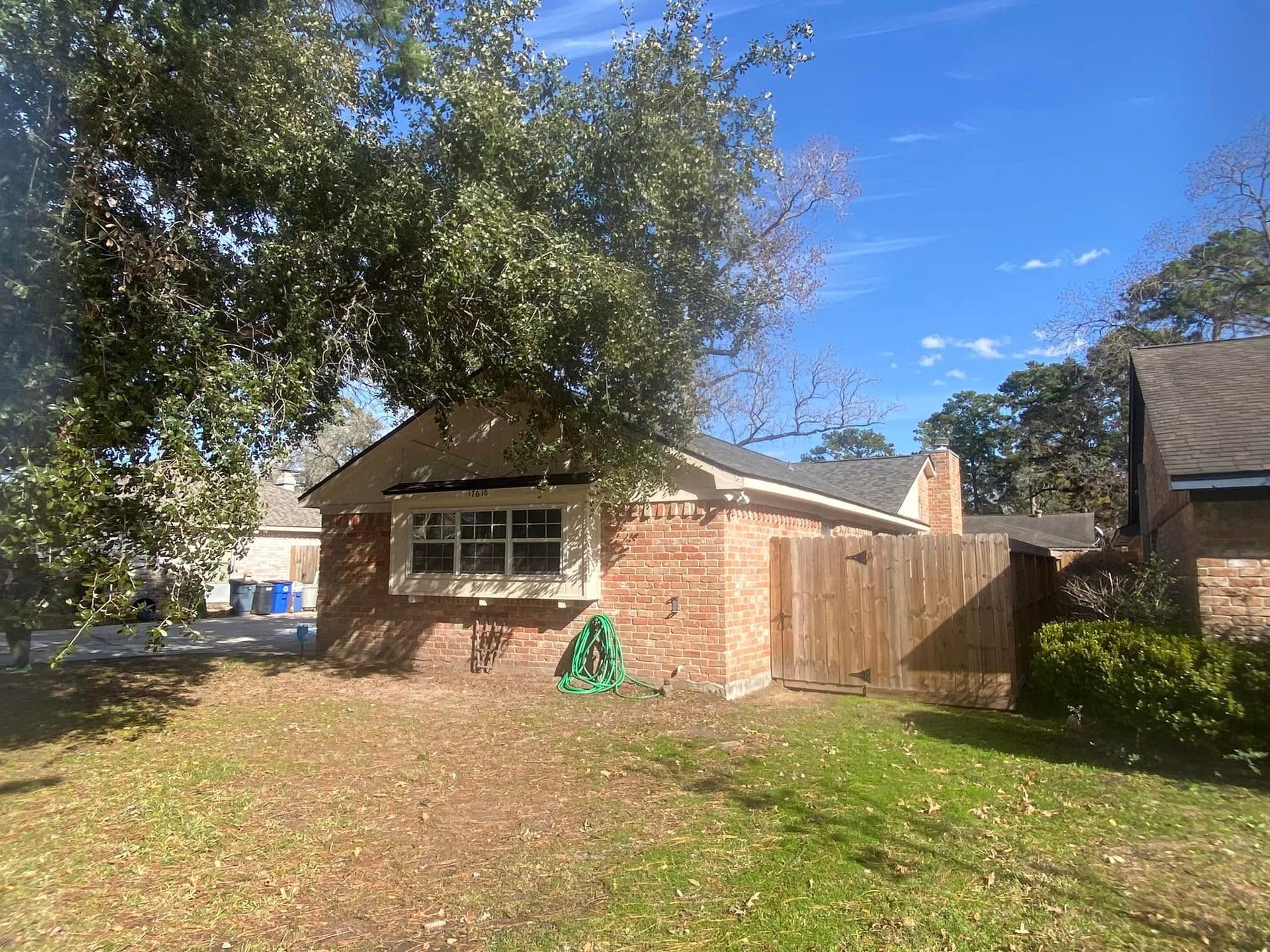 Brick house with window, wooden fence, tree, green hose, and brown grass on a sunny day.