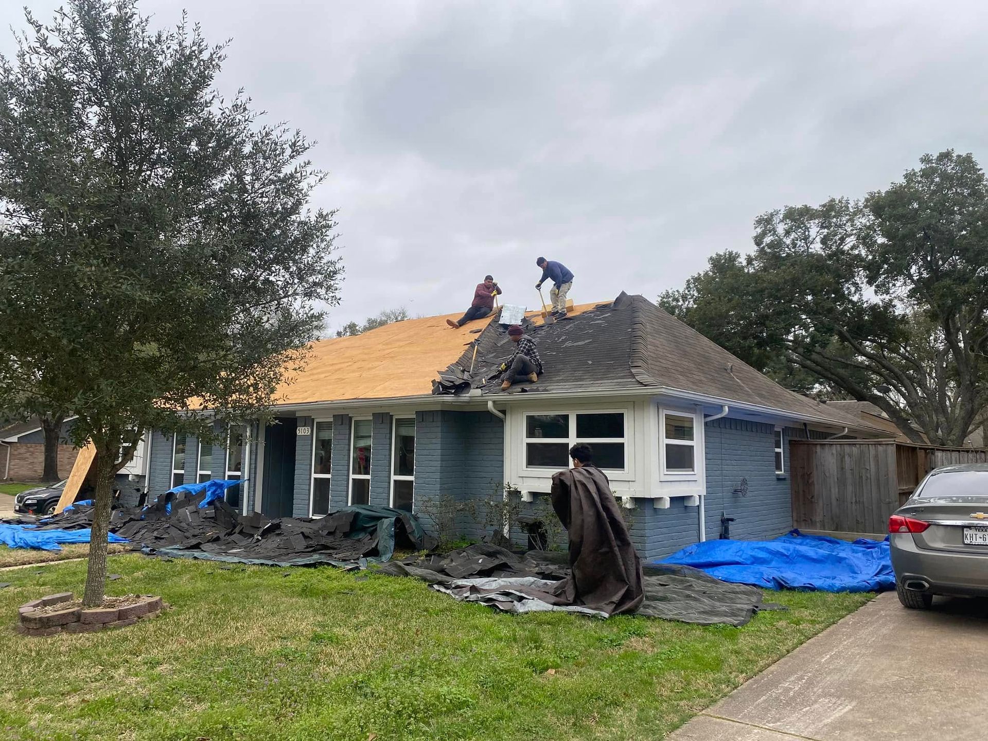 Workers removing old shingles and replacing a roof on a blue house under an overcast sky.