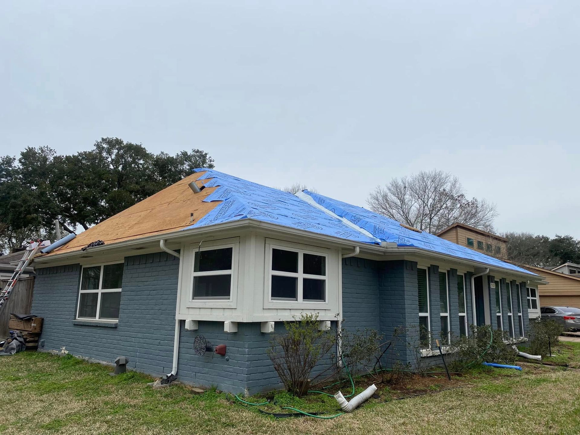 House with a partially replaced roof; blue tarp covering part of it. Brick exterior, white-framed windows, cloudy sky.