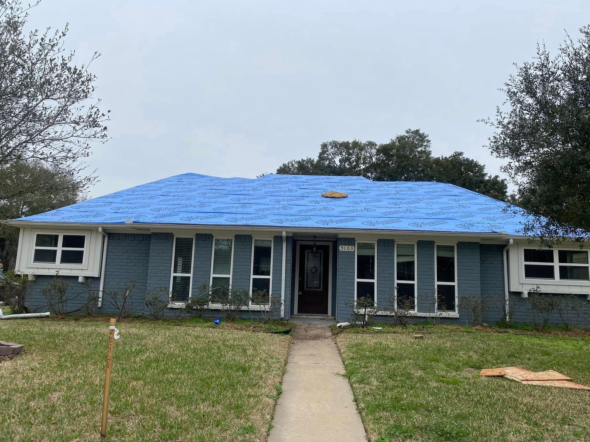 House with blue tarp roof; weathered blue brick exterior, white window trim, cloudy sky.