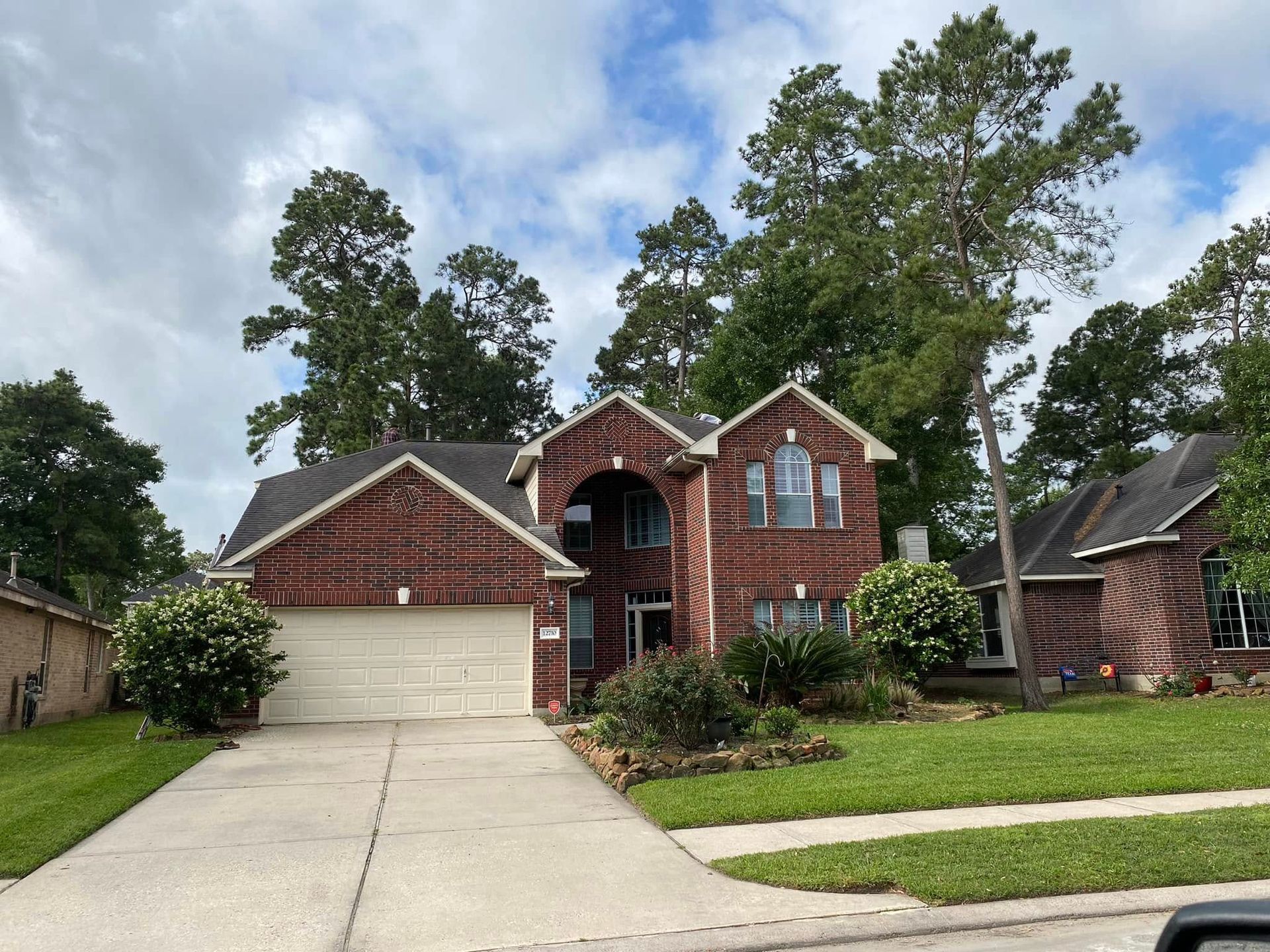 Two-story brick house with attached garage, green lawn, trees, and cloudy sky.