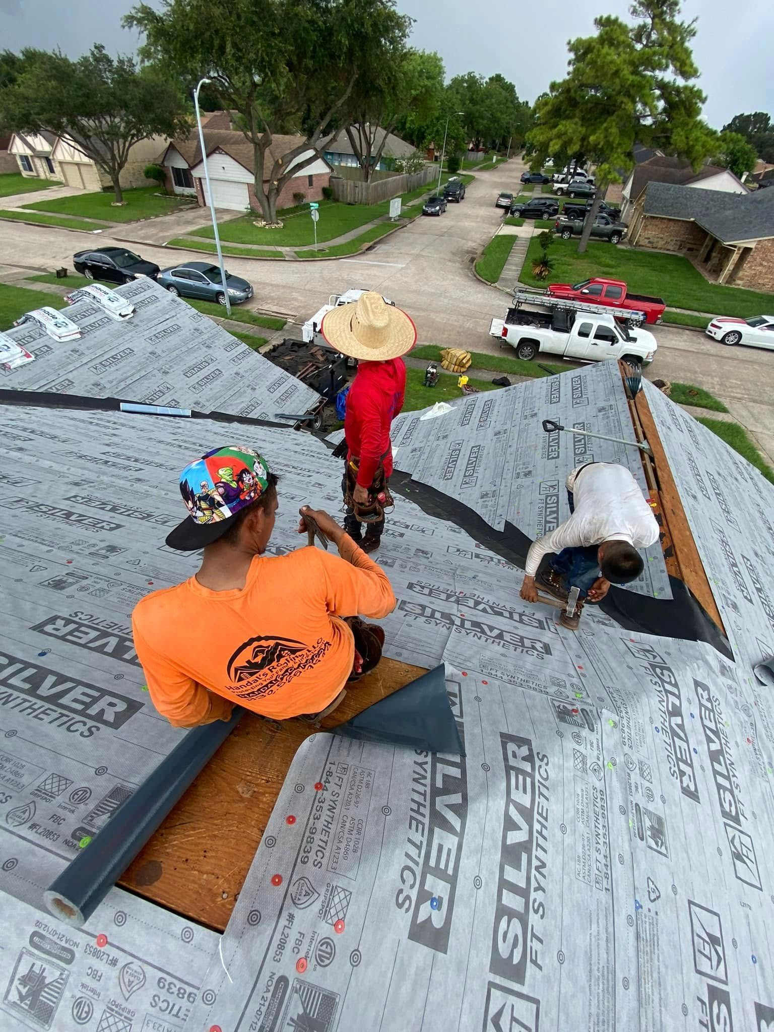 Three roofers installing shingles on a house roof. One wears an orange shirt and cap, two others wear a hat and white shirt.