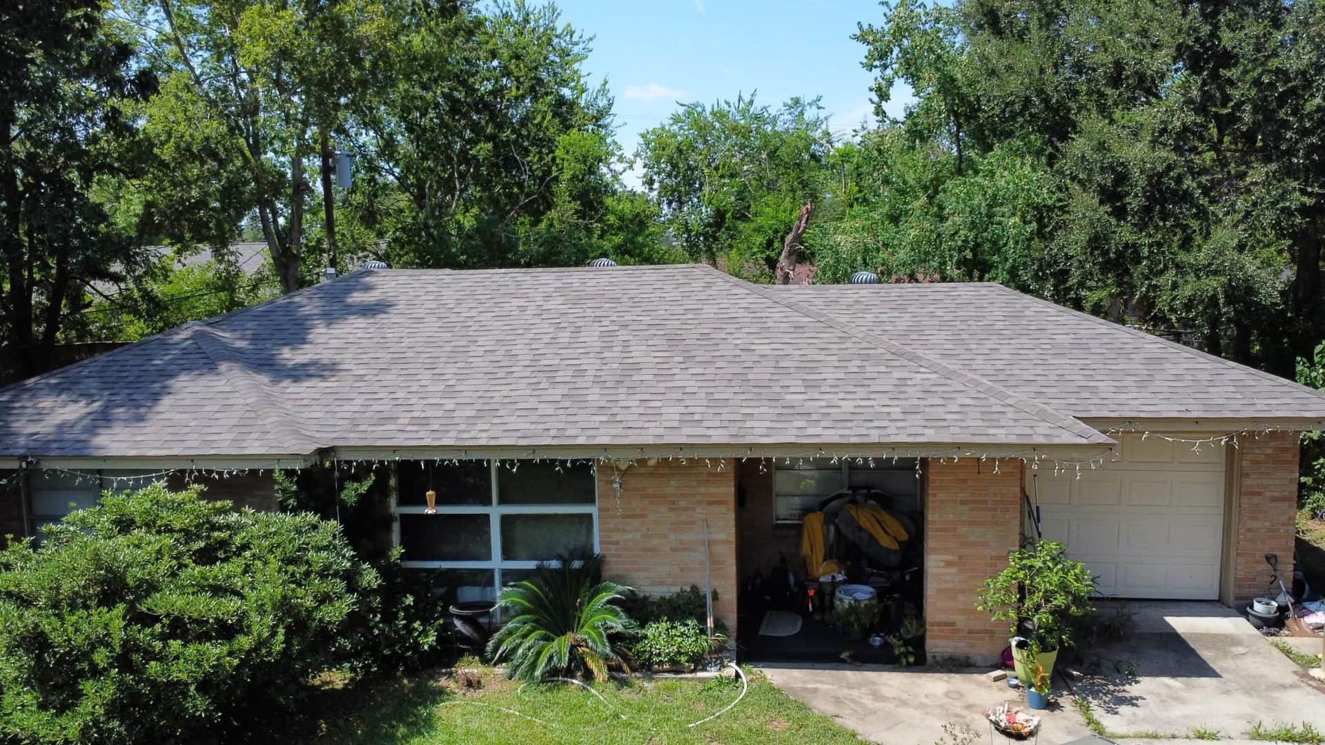 House with brown roof and brick exterior, surrounded by green trees and foliage.