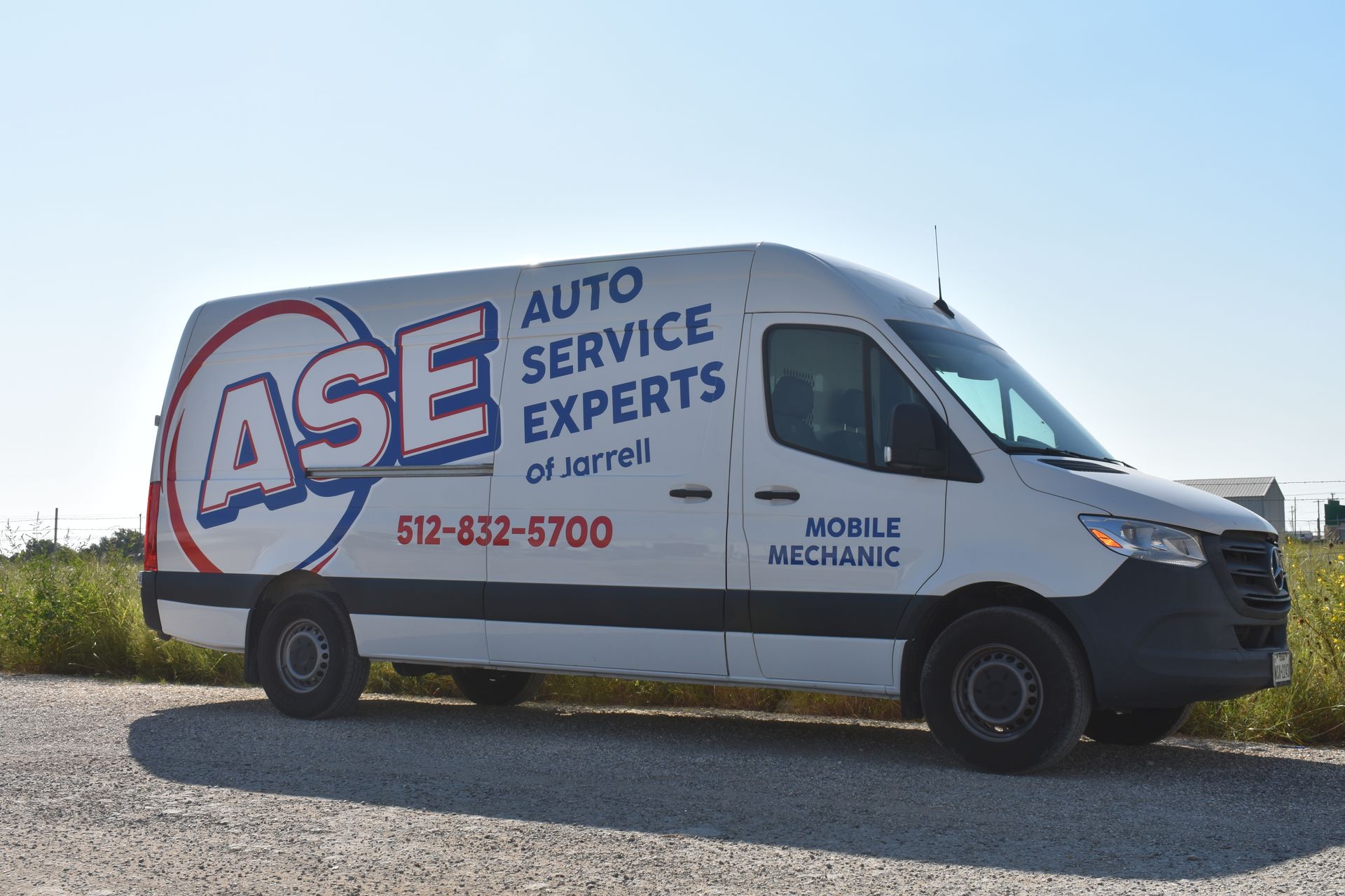 White ASE auto service van with logo parked on a gravel road under a blue sky.