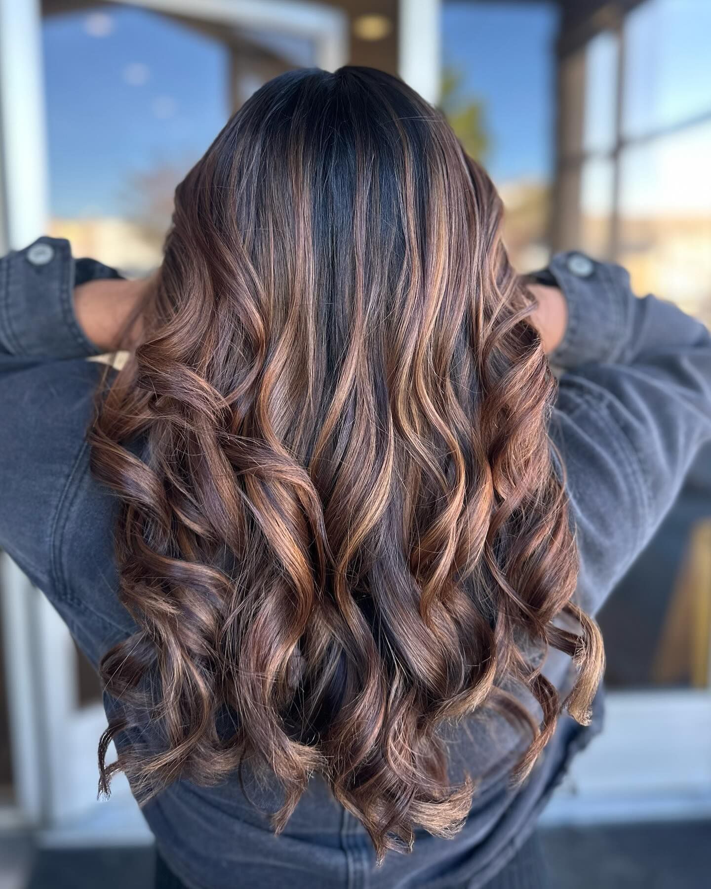 A woman with long brown hair is standing in front of a window with her hands behind her head.