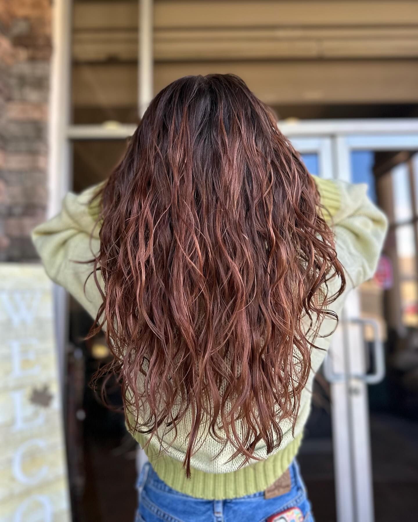 A woman with long curly hair is standing in front of a door with her hands behind her head.