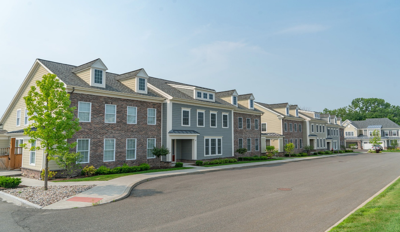 Row of townhouses with varying color brick and siding, sidewalk, and street under a blue sky.