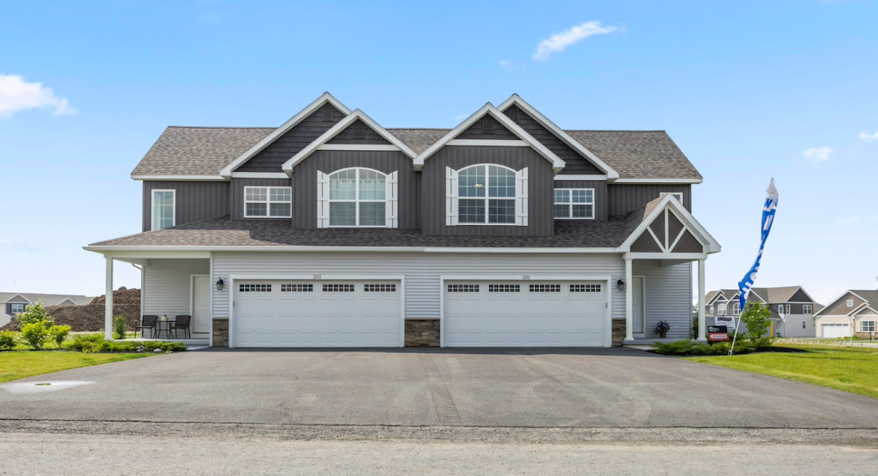 Two-story duplex with gray siding, white trim, and two-car garages on a paved driveway under a blue sky.