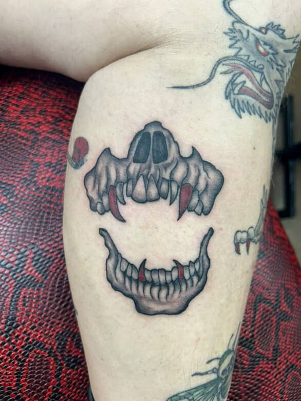Skull tattoo on leg: grey-shaded animal skull with sharp teeth and jawline, red accents.