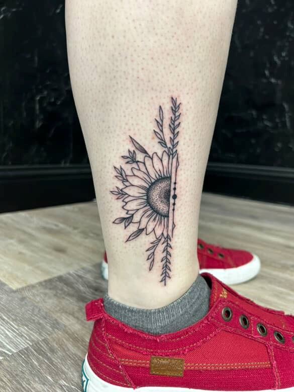 Tattoo of a sunflower and leaves on a leg, next to a person's red shoe.