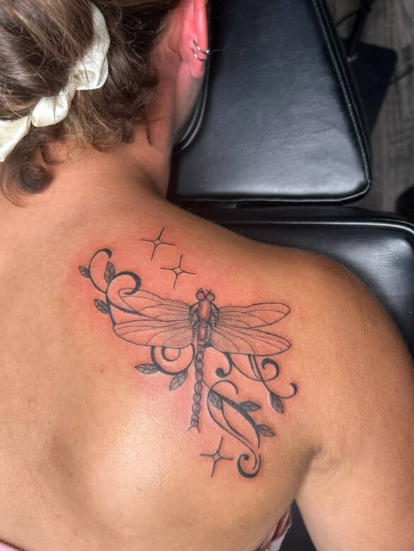 Woman's back with a dragonfly tattoo. Black ink outlines with stars and foliage.