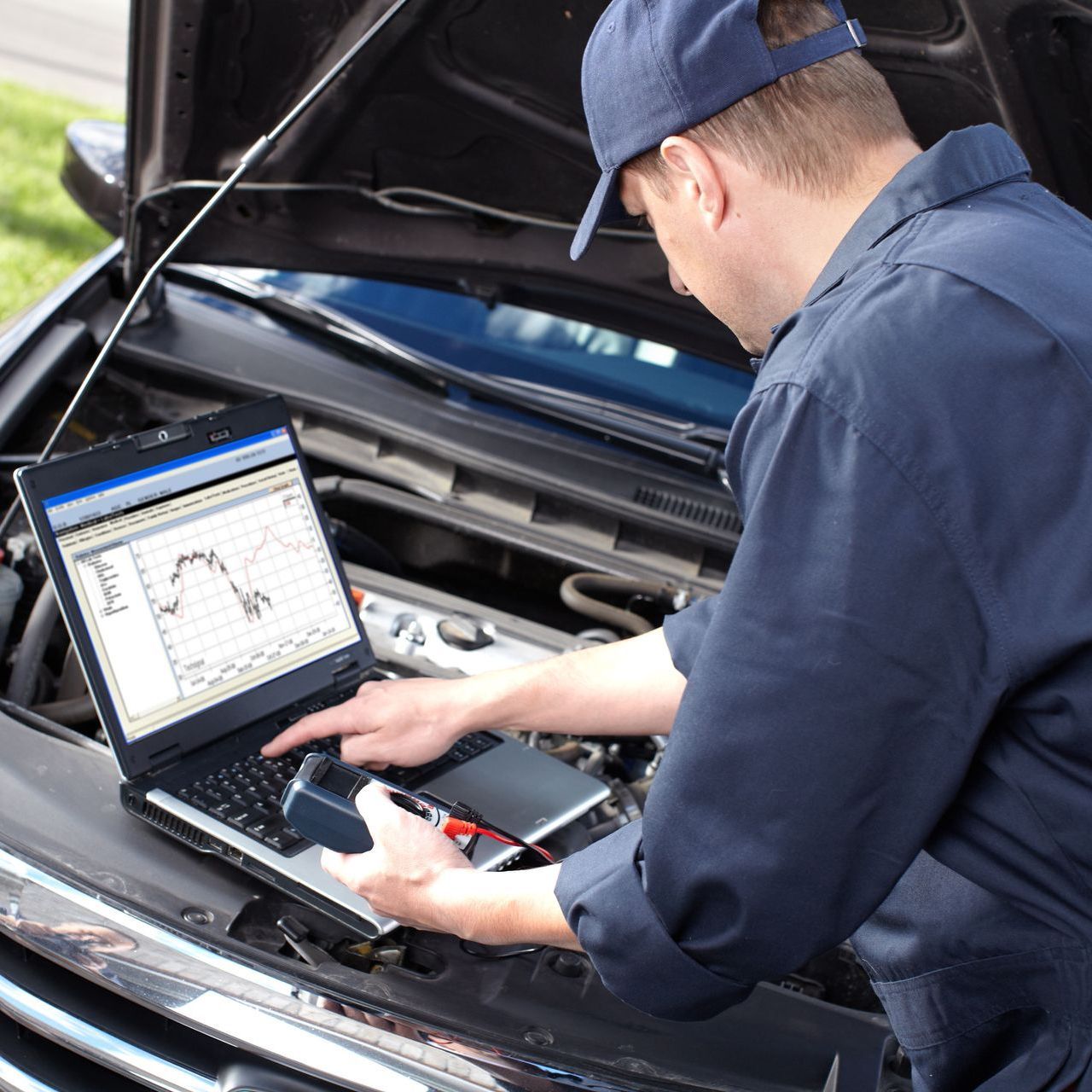 A man is working on a laptop under the hood of a car