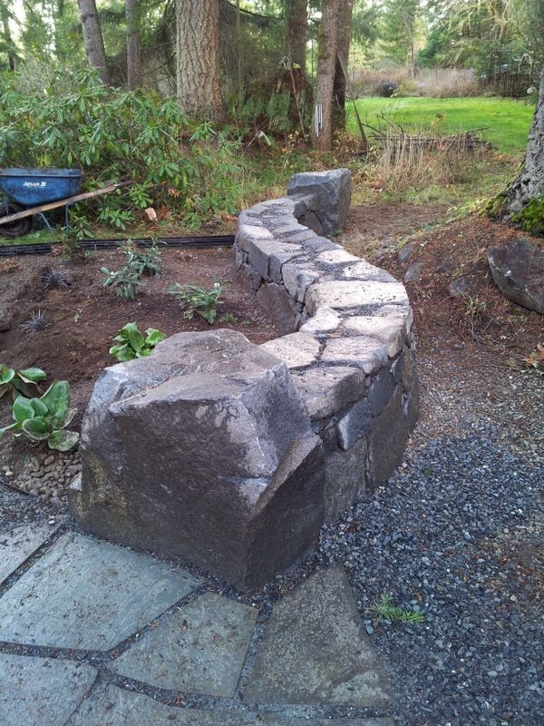 A stone wall is being built in a garden with a blue wheelbarrow in the background.