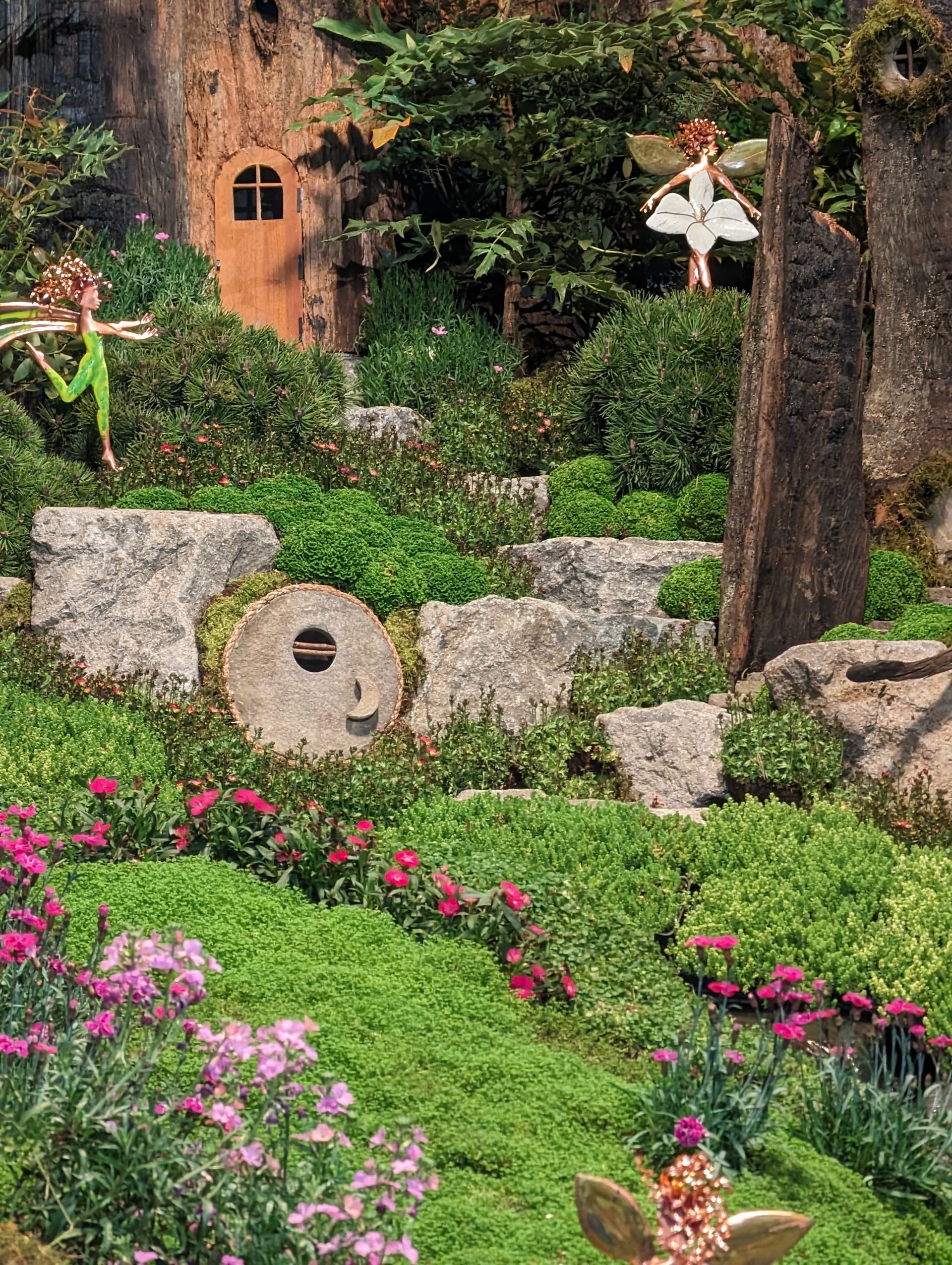 A fairy garden with flowers , rocks , trees and a house in the background.
