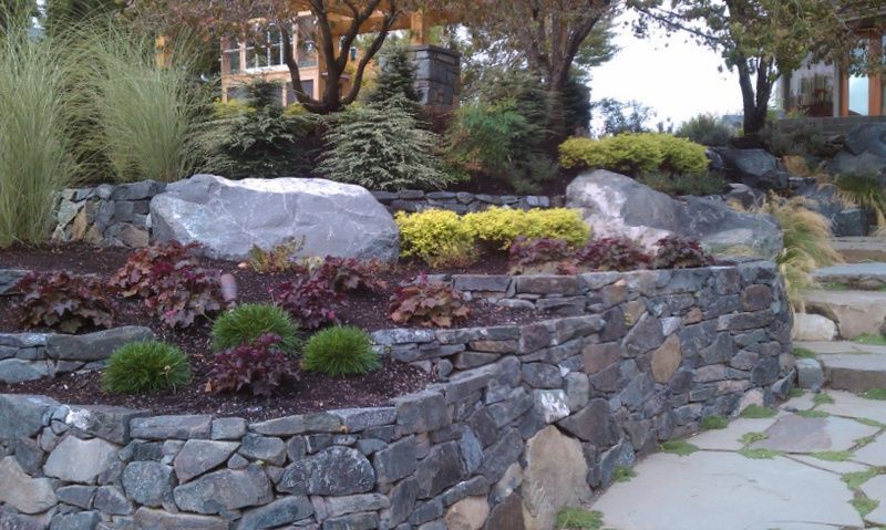 a stone wall is surrounded by plants and rocks in a garden