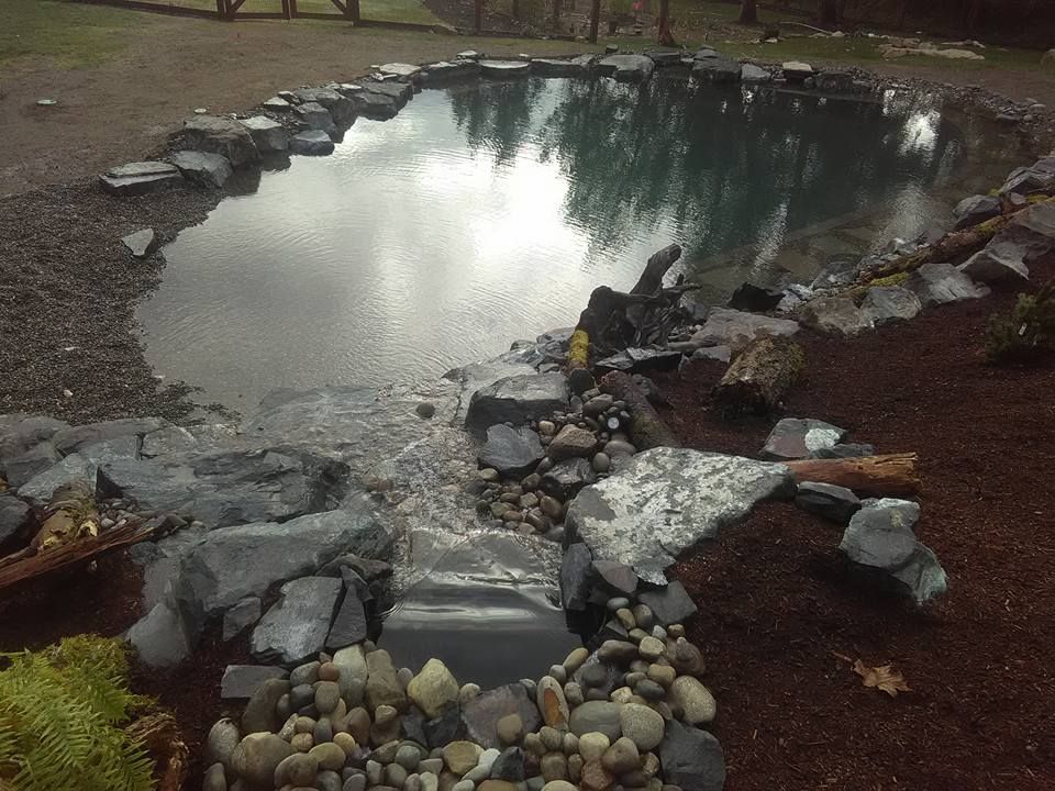 a pond surrounded by rocks and gravel with trees reflected in it