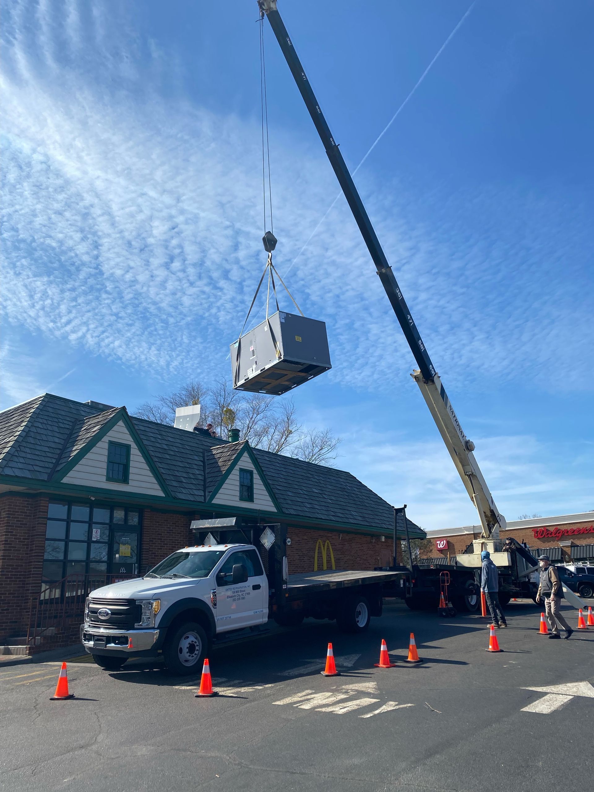 A crane lifts a large HVAC unit above a building. A truck and cones are on the pavement. Sunny, blue sky.