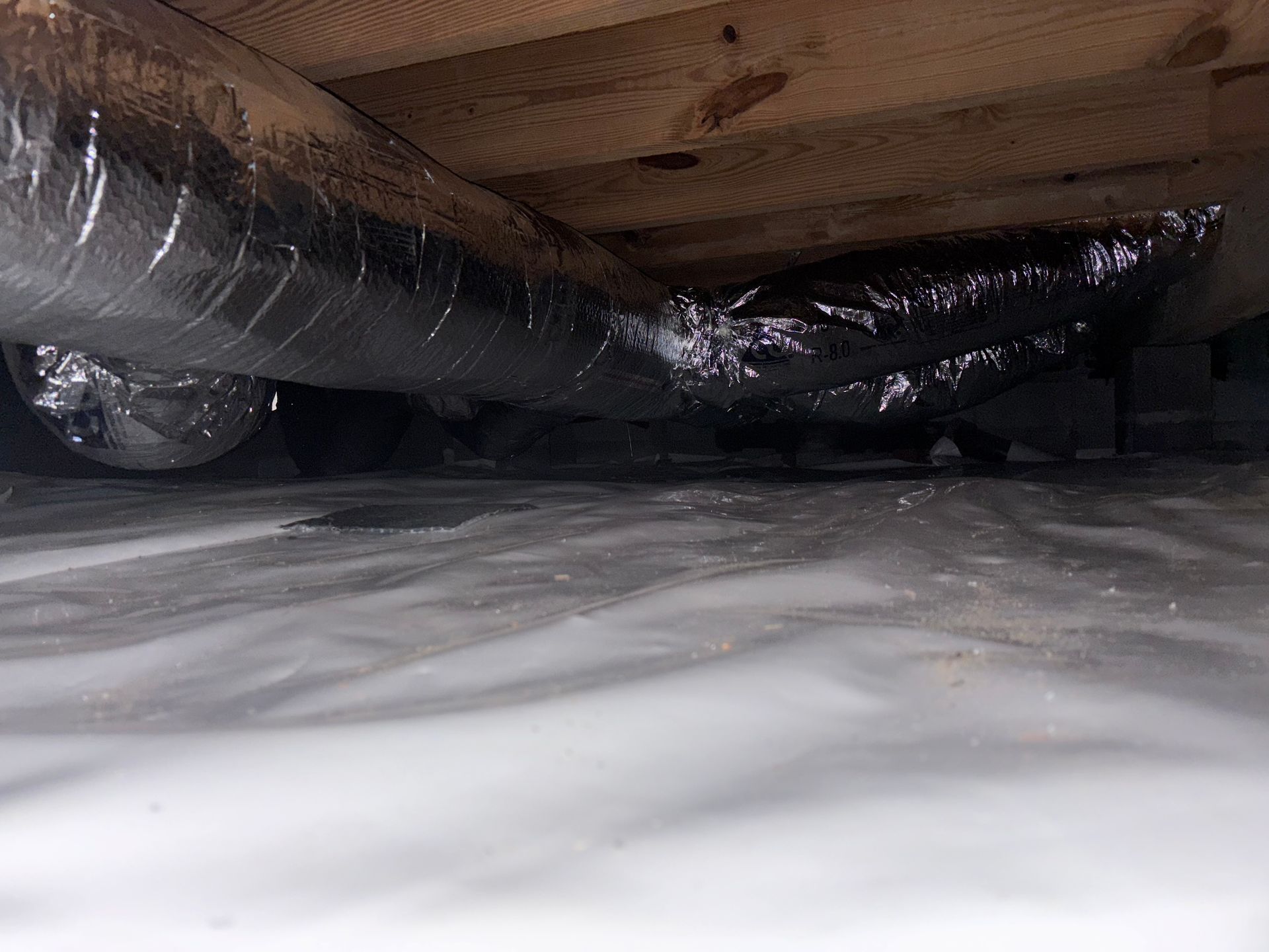 View of ductwork and plastic sheeting in a crawl space under wooden beams.