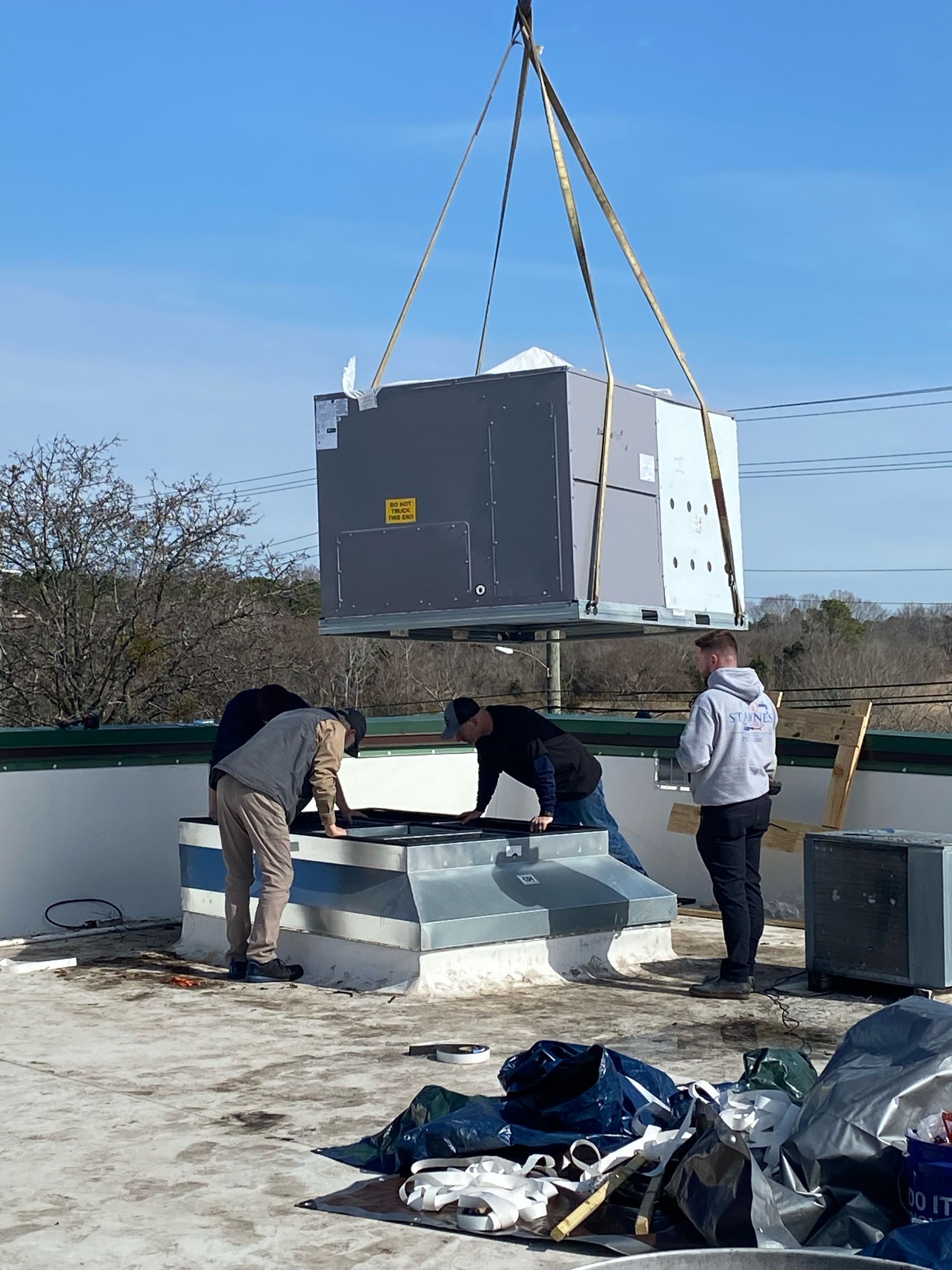 A crane lifts HVAC unit above a roof as three workers prepare for installation.