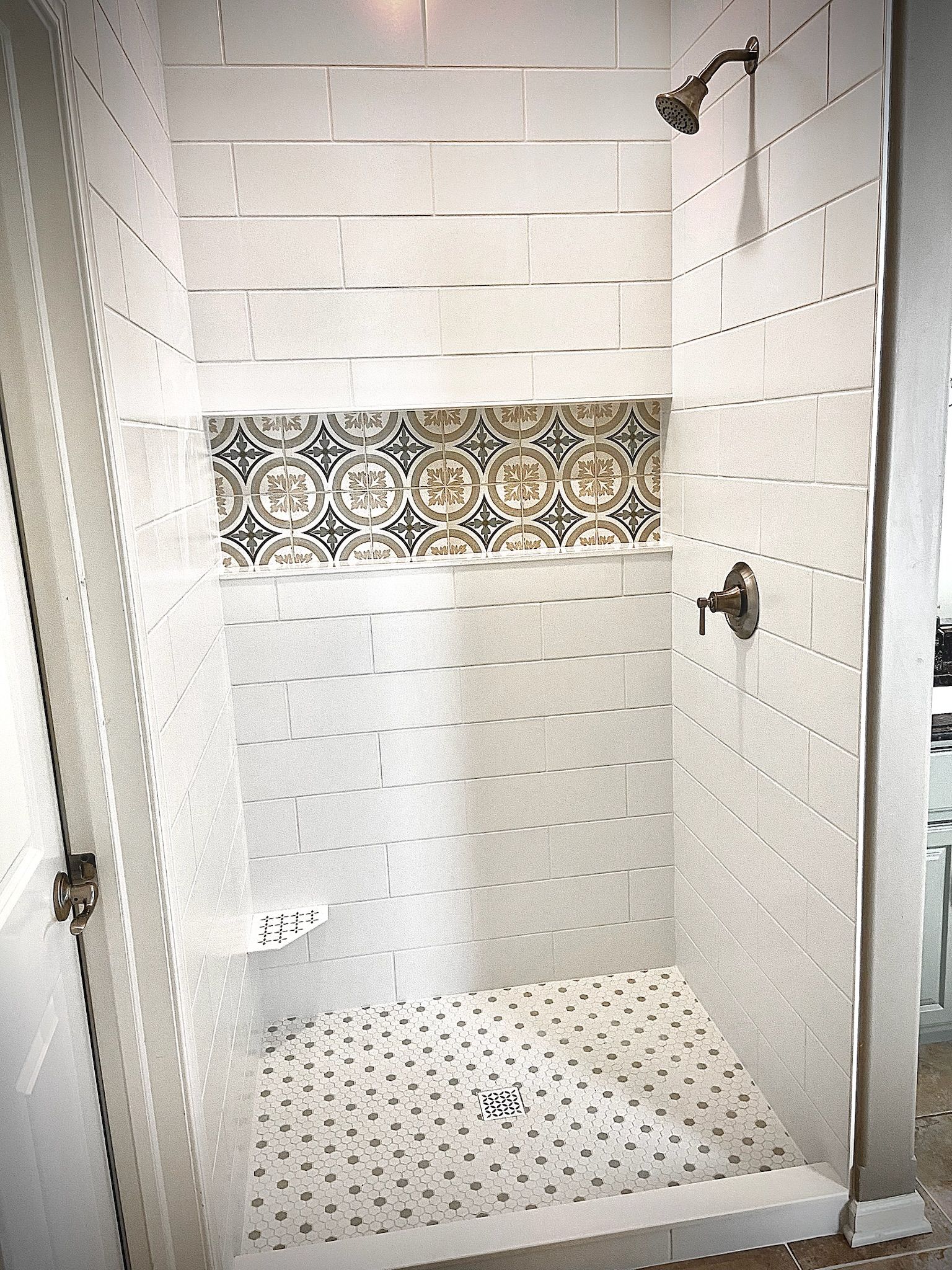 White tiled shower with decorative tile accent and pebble-style floor. Bronze fixtures.