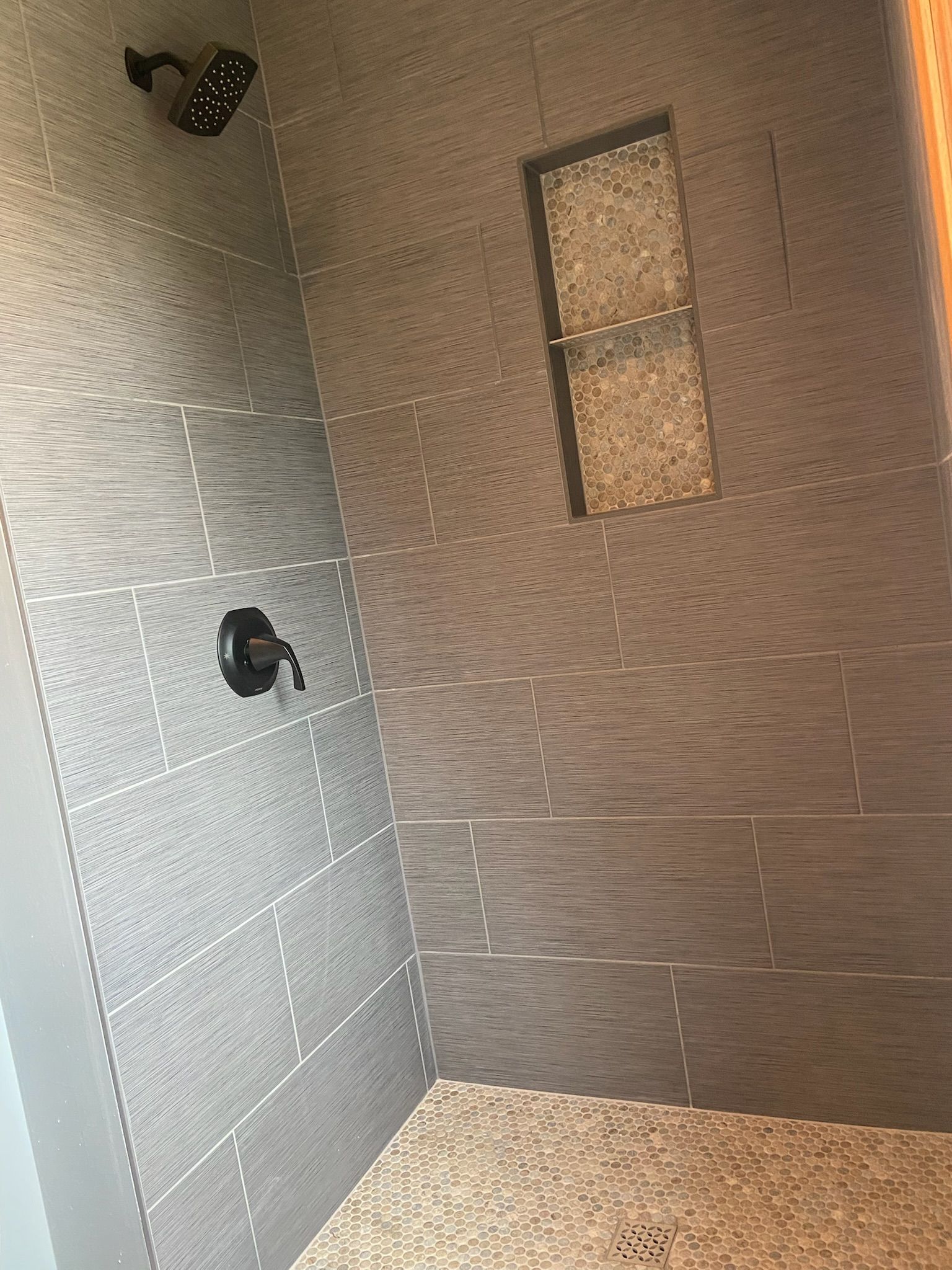 Grey tiled shower with pebble floor, built-in shelf, and black shower fixtures.