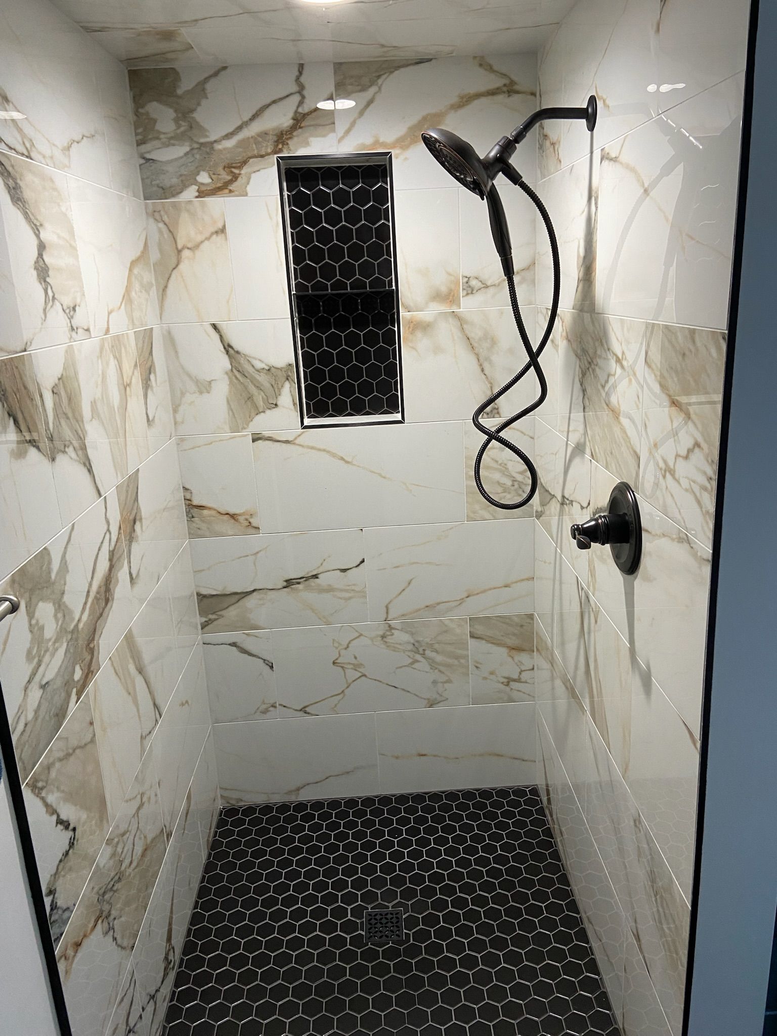 Shower with marble-patterned tile walls, a black pebble floor, and a black showerhead.