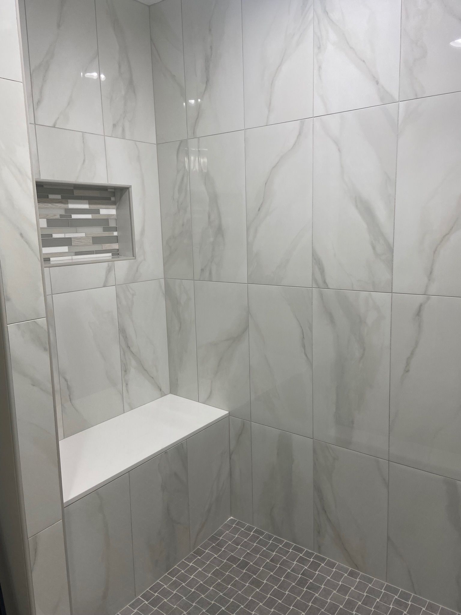 Shower with white marble-look tiles, built-in shelf, and recessed niche. Gray stone tile floor.