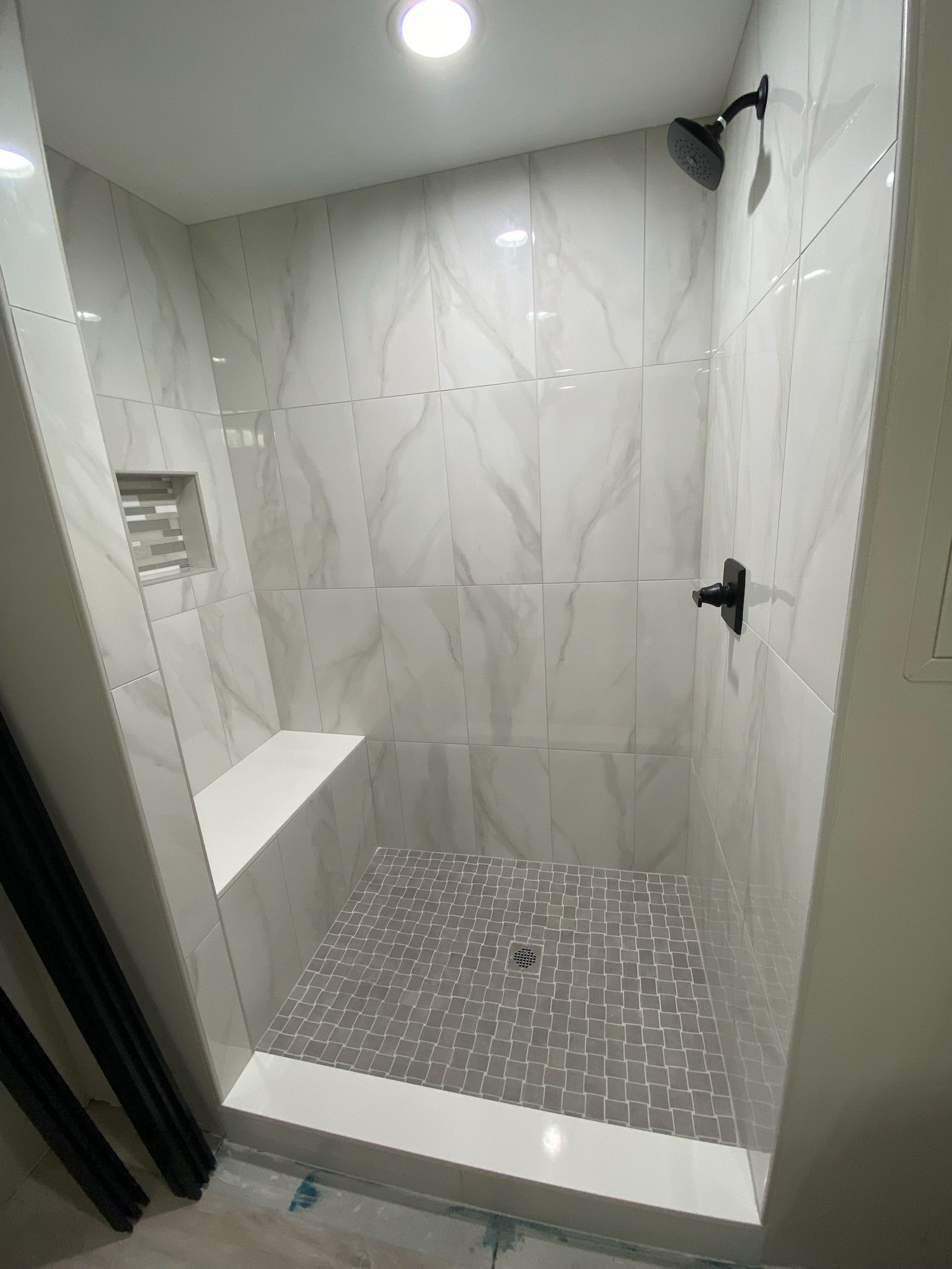 Modern bathroom shower with white marble-like tile walls and a grey tile floor. Includes a built-in bench.