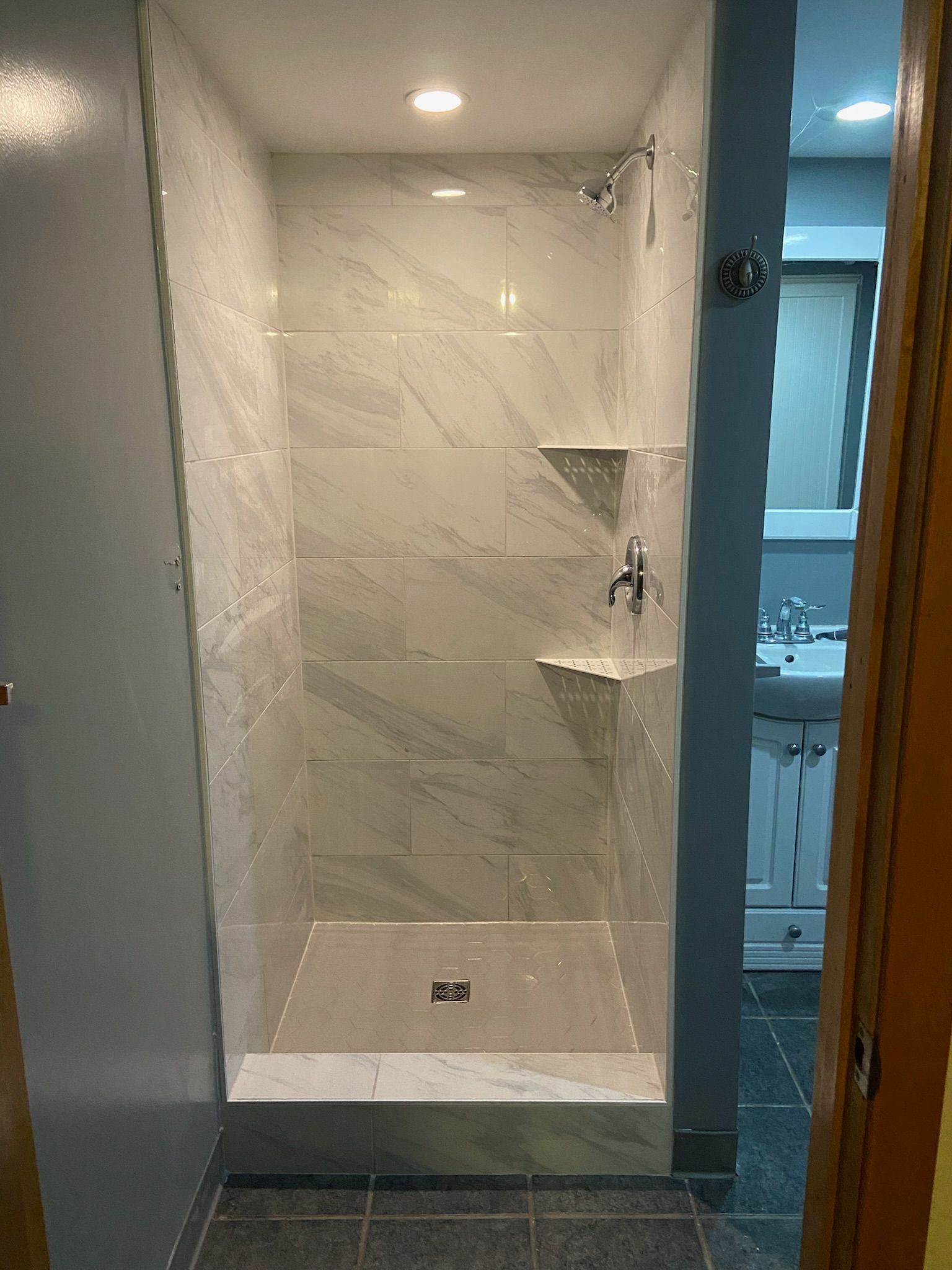 Shower stall with white marble-like tiles and chrome fixtures; doorway on the left, bathroom visible to the right.