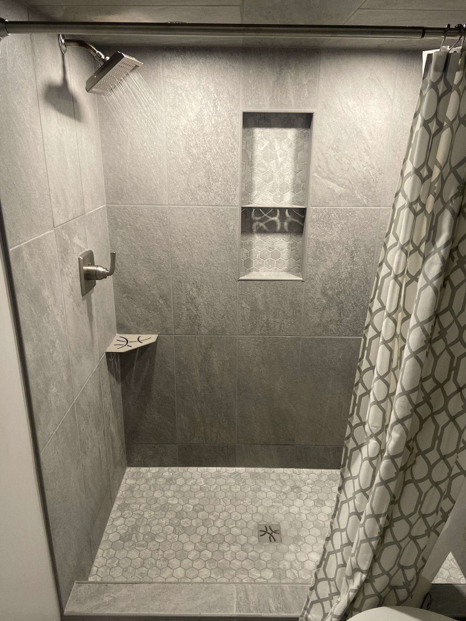Gray tile shower with showerhead, built-in shelves, and patterned shower curtain.