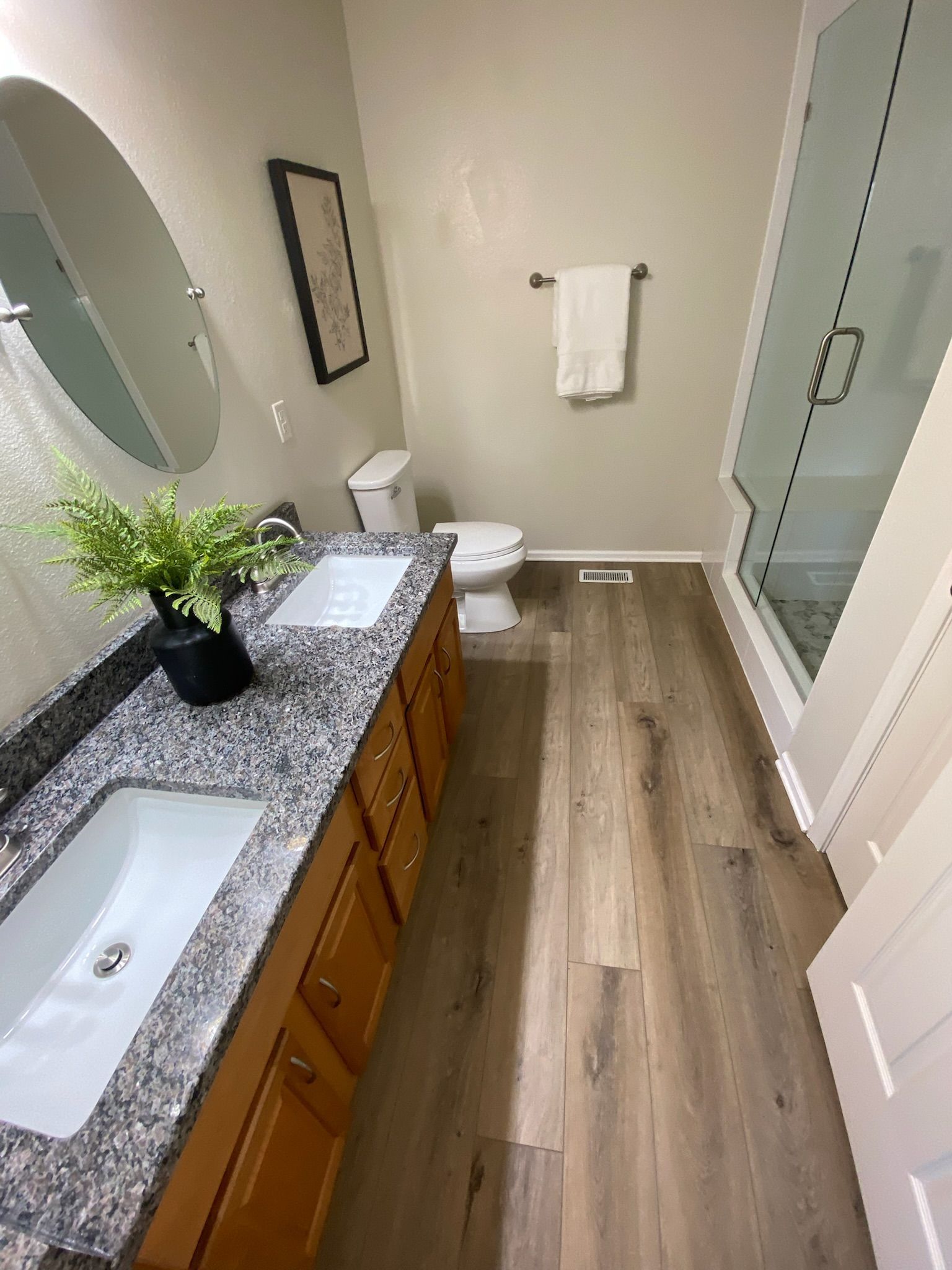 Bathroom with double vanity, granite countertop, wooden cabinets, shower, toilet, and wood-look flooring.